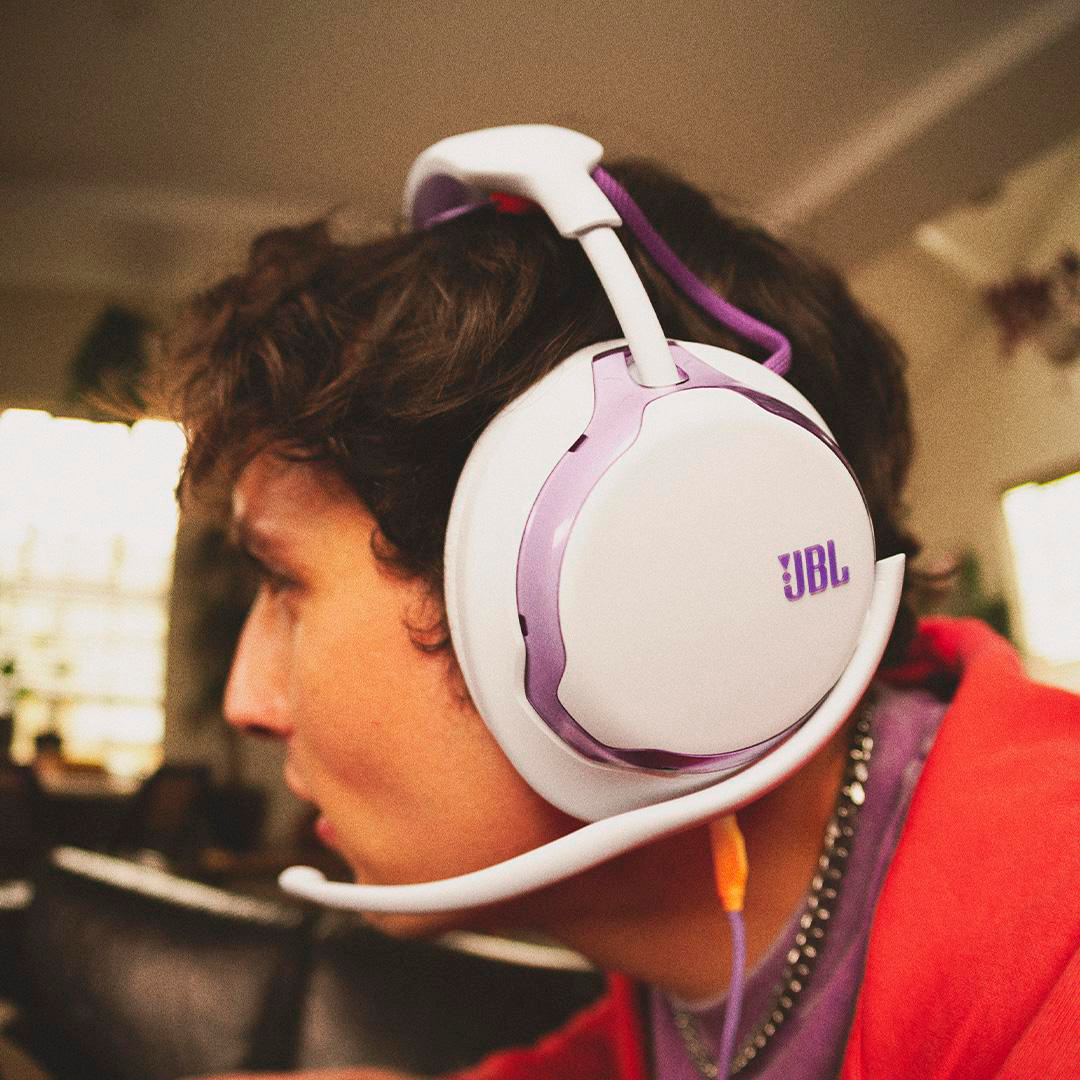 Eine Person trägt ein weißes und lila JBL-Headset. Sie trägt eine rote Jacke und schaut nach links. Ein Mikrofon ist sichtbar.