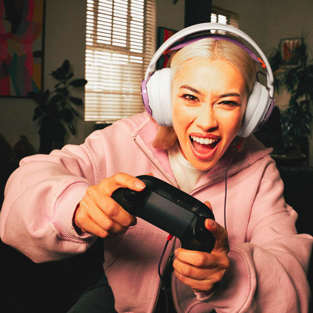 Blonde Frau mit Kopfhörern spielt Videospiel, hält Controller, lächelt breit und trägt einen rosa Hoodie.