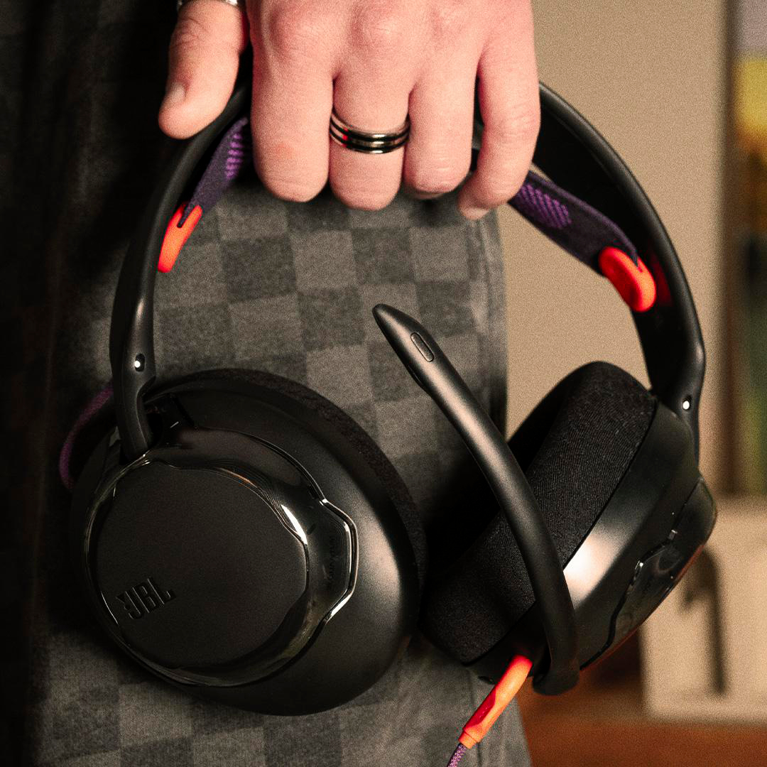 Eine Person hält schwarze JBL-Kopfhörer. Die Kopfhörer haben lila und orangefarbene Akzente. Die Person trägt einen Ring. Der Hintergrund ist grau.
