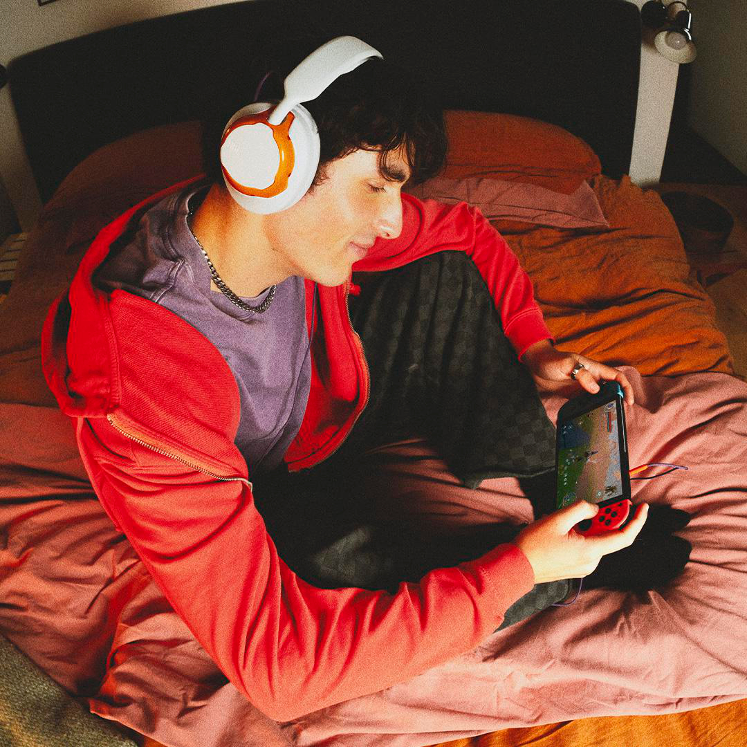Eine Person mit Kopfhörern und roter Jacke spielt ein Handheld-Spielgerät, während sie auf einem Bett mit roten Laken sitzt.