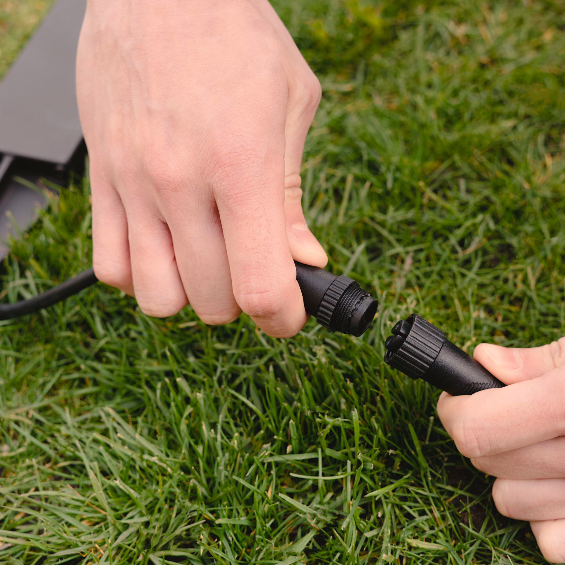 Hände verbinden einen schwarzen Kabelstecker vor grünem Gras. Die Hände sind hellhäutig.
