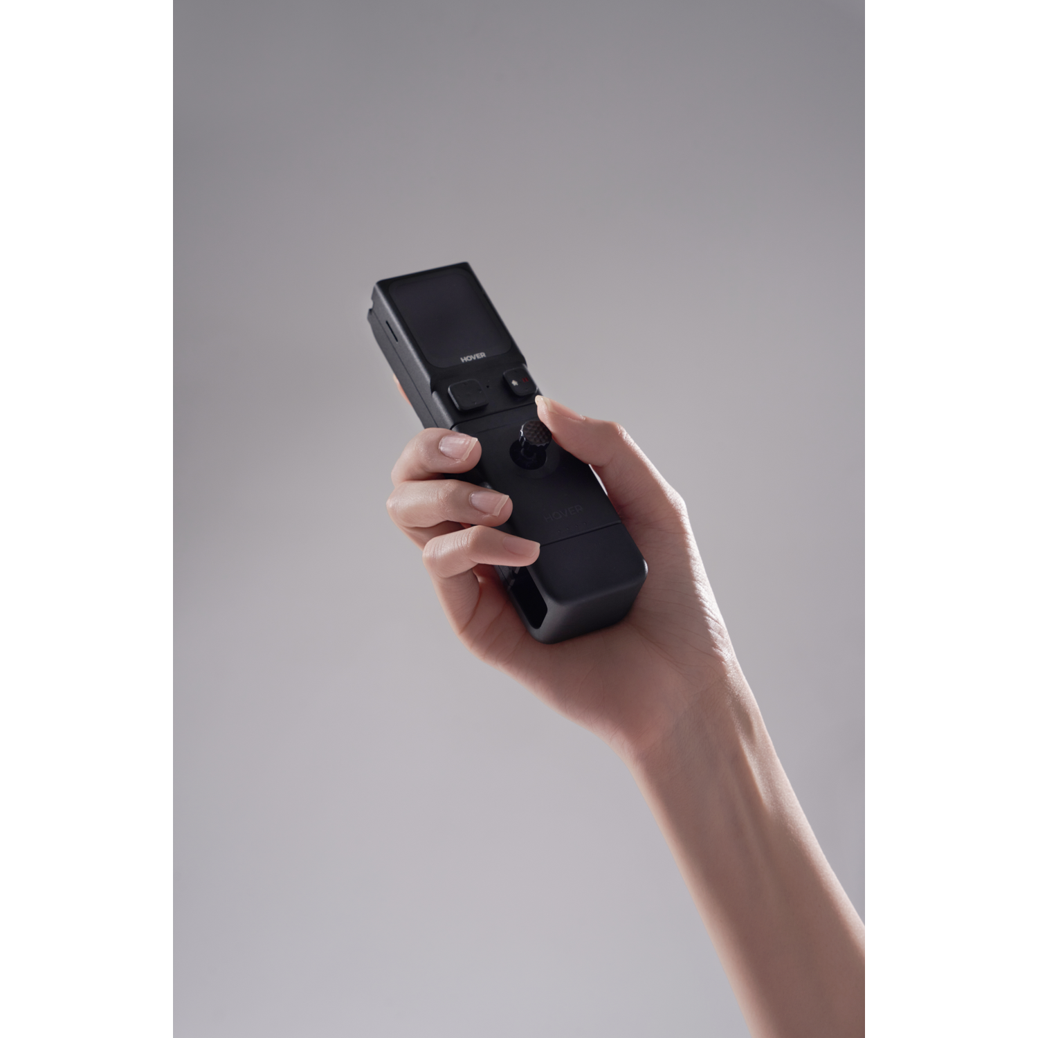 Een hand die een zwart apparaat met een scherm en een joystick vasthoudt. De achtergrond is effen grijs.