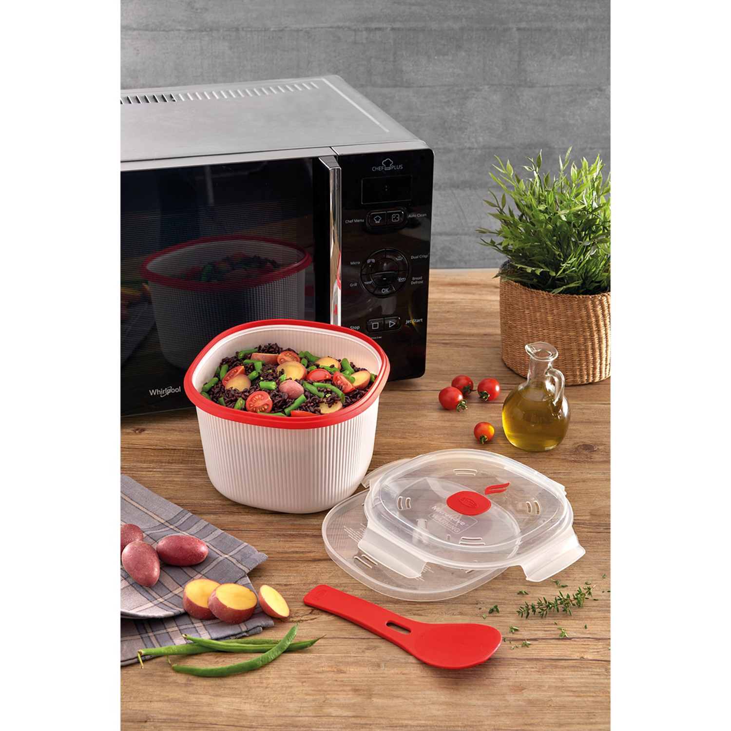 Microonde, ciotola di cibo, verdure, pomodori, bottiglia d'olio e una pianta su un tavolo di legno.