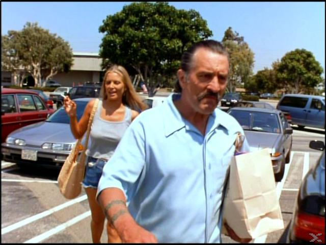 Ein Mann im blauen Hemd trägt eine Tüte. Eine Frau in Jeansshorts und einem Tanktop geht mit ihm in der Nähe von Autos.