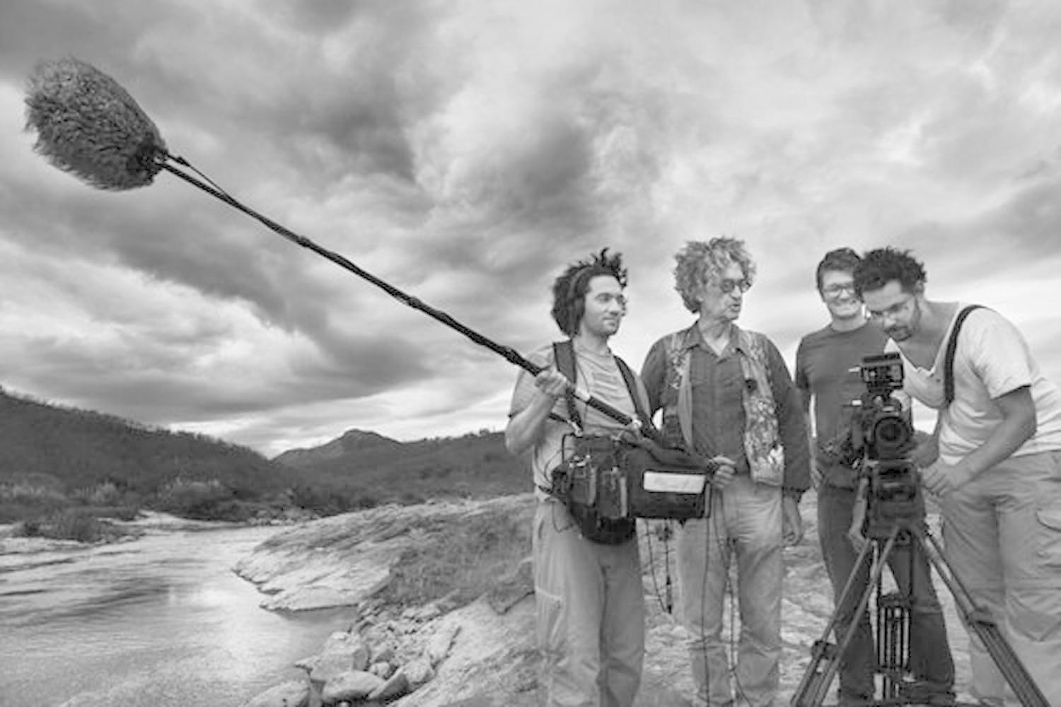 Filmteam vor Ort. Ein Mann hält eine Mikrofonangel. Andere betrachten eine Kamera auf einem Stativ. Fluss und Hügel im Hintergrund.