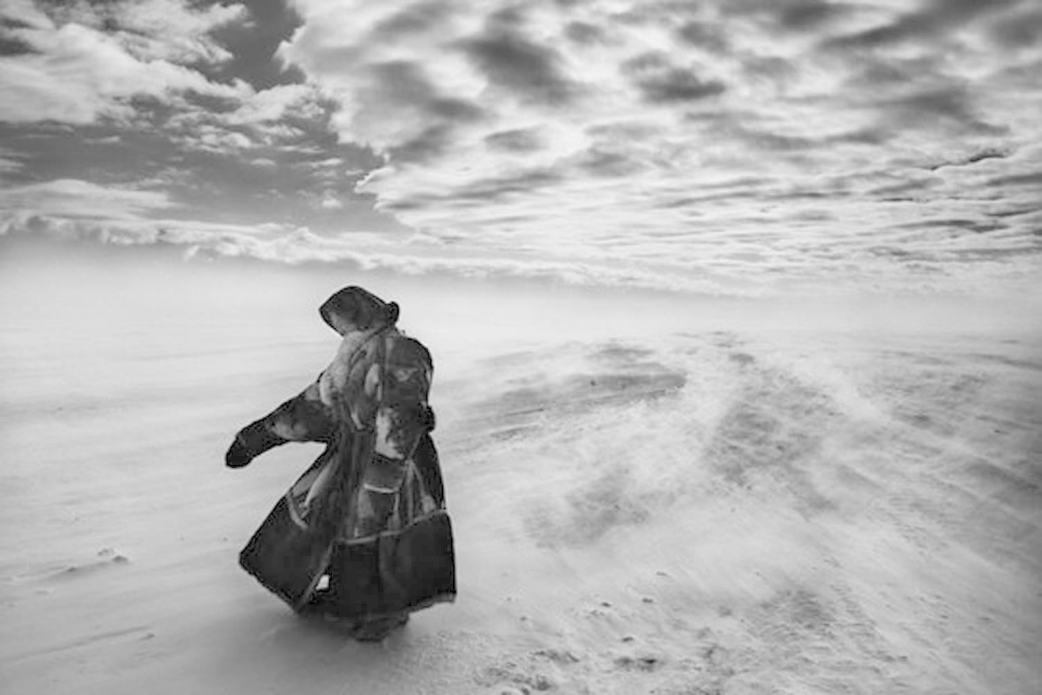 Eine Person in einem Mantel geht im Schnee, Wind weht. Bewölkter Himmel im Hintergrund. Schwarz-Weiß-Bild.