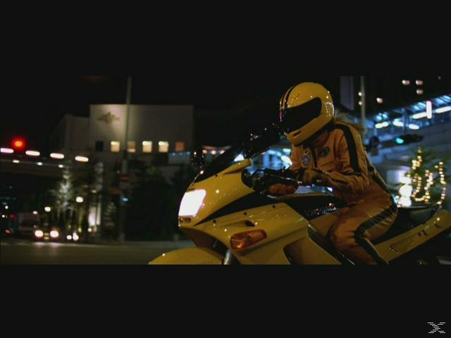 Eine Person in gelbem Motorradanzug und Helm fährt nachts auf einem gelben Motorrad. Stadtlichter sind im Hintergrund zu sehen.
