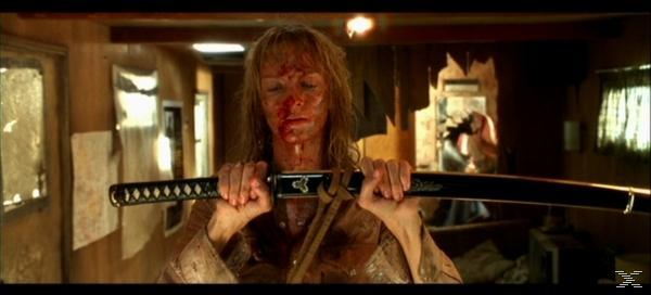 Eine Frau mit Blut im Gesicht hält ein Katana. Sie befindet sich in einer Holzkonstruktion. Im Hintergrund ist eine Person zu sehen.