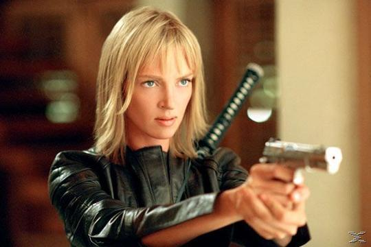 Blonde Frau hält eine Pistole, trägt eine schwarze Jacke und hat ein Schwert auf dem Rücken. Sie blickt aufmerksam.