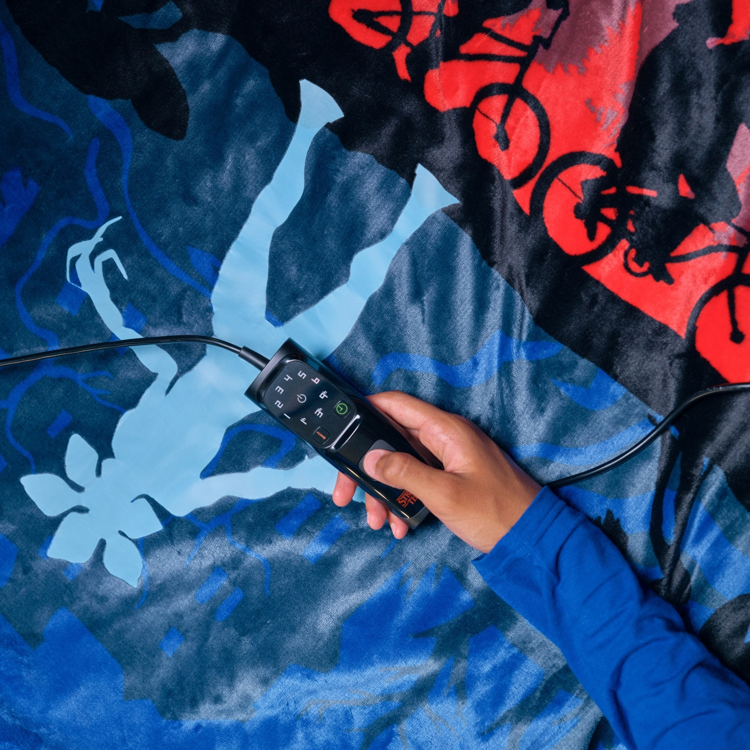 Una mano tiene un telecomando nero su una coperta. La coperta ha immagini di biciclette, un fiore e una persona. Manica blu visibile.