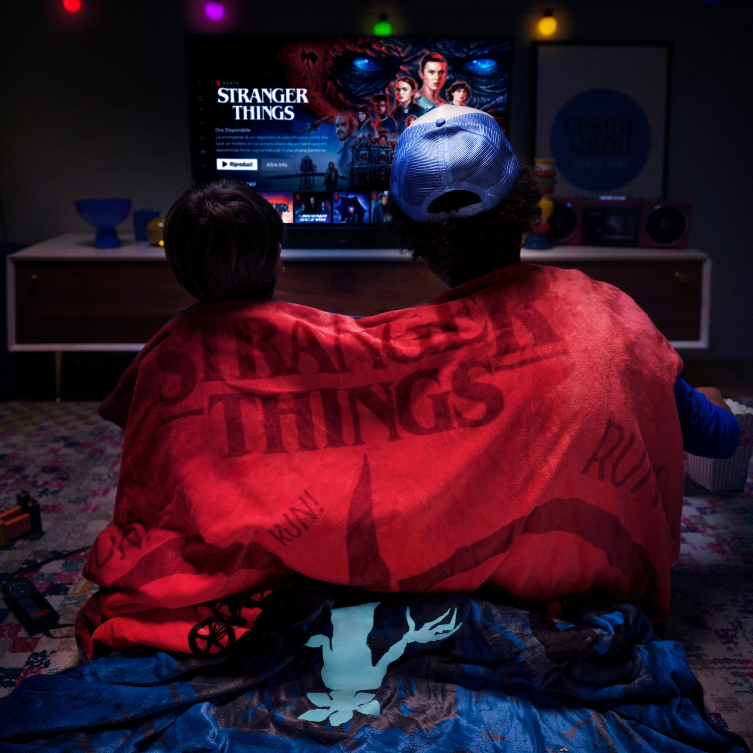 Due persone guardano la TV, coperte da una coperta rossa con "Stranger Things". La TV mostra lo stesso titolo. Le luci sono accese.