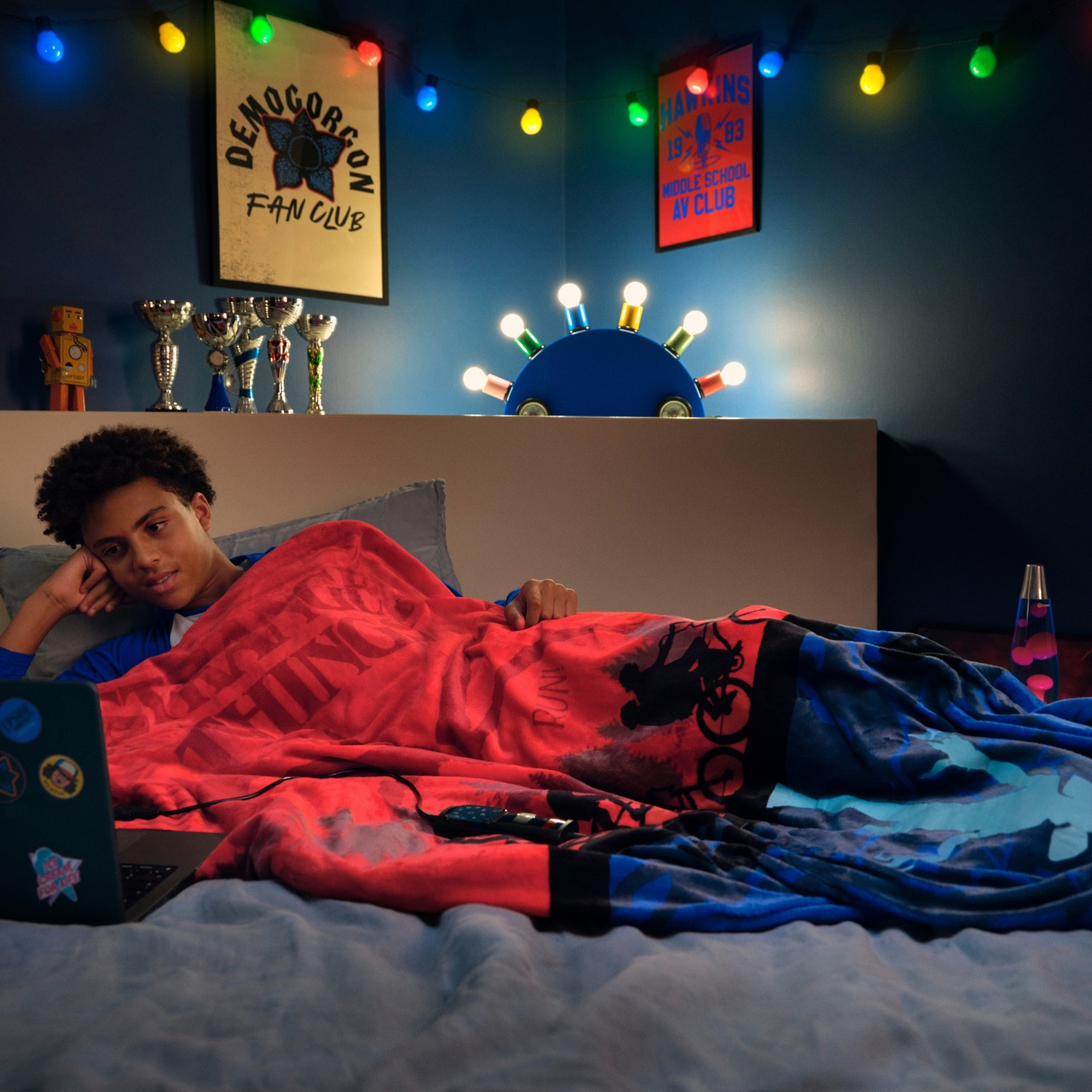 Adolescente a letto, guarda il laptop. Coperta rossa con logo della serie. Luci a corda, trofei e poster sullo sfondo.