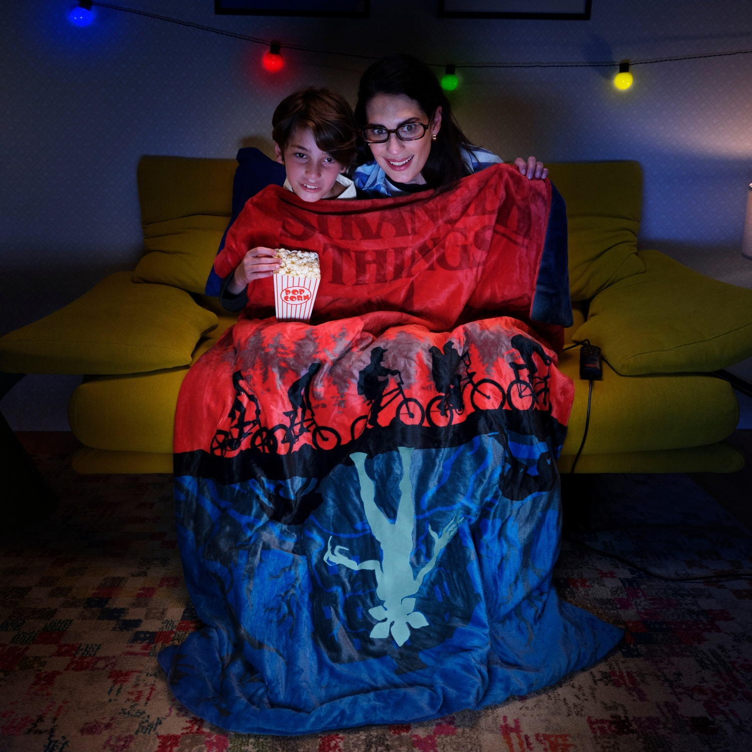 Una donna e un ragazzo su un divano, coperti da una coperta. Tengono popcorn. Luci a fatina sopra. La coperta ha un disegno.