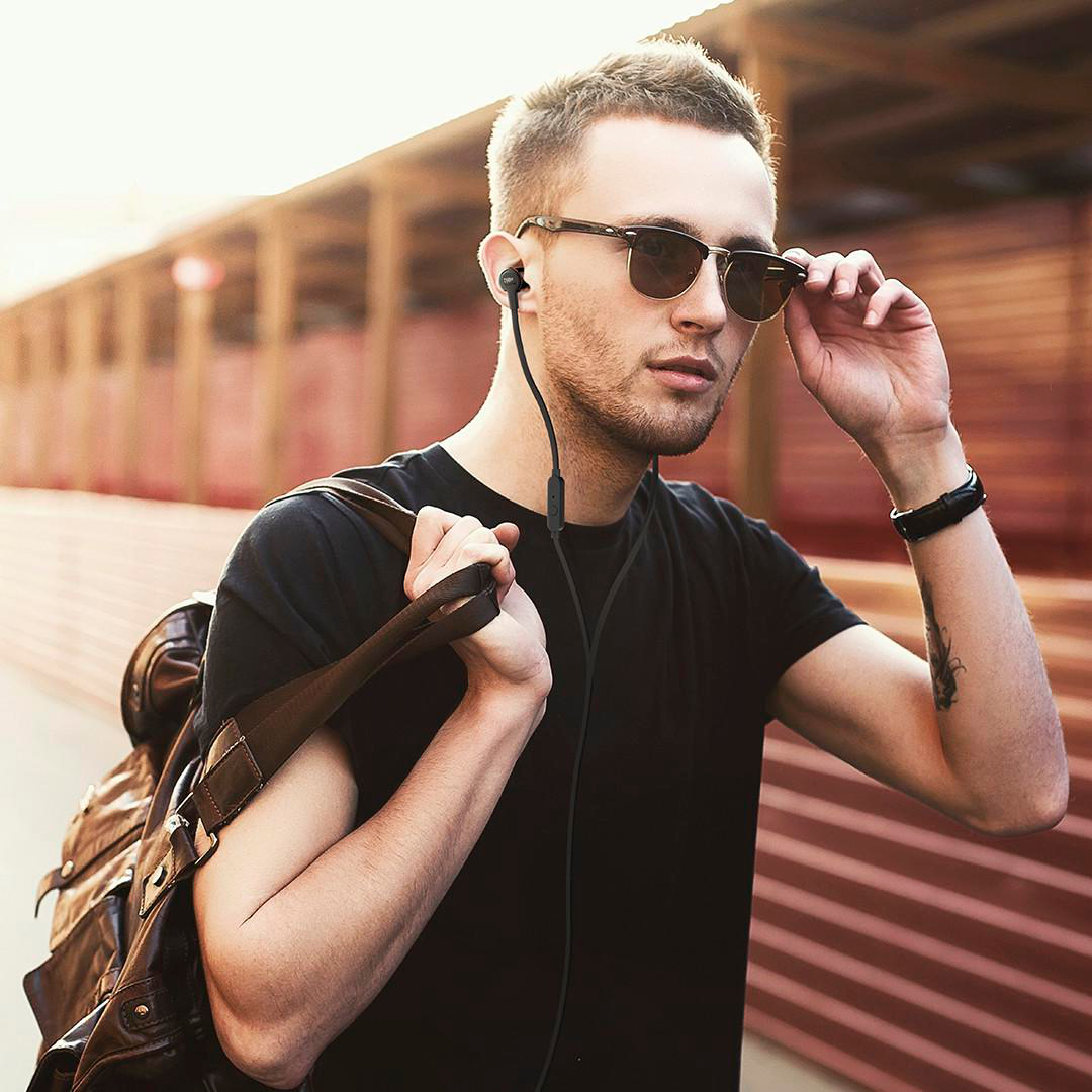 Ein Mann mit Sonnenbrille und Kopfhörern, der eine Tasche hält und seine Brille richtet. Er trägt ein schwarzes Hemd und hat ein Tattoo.