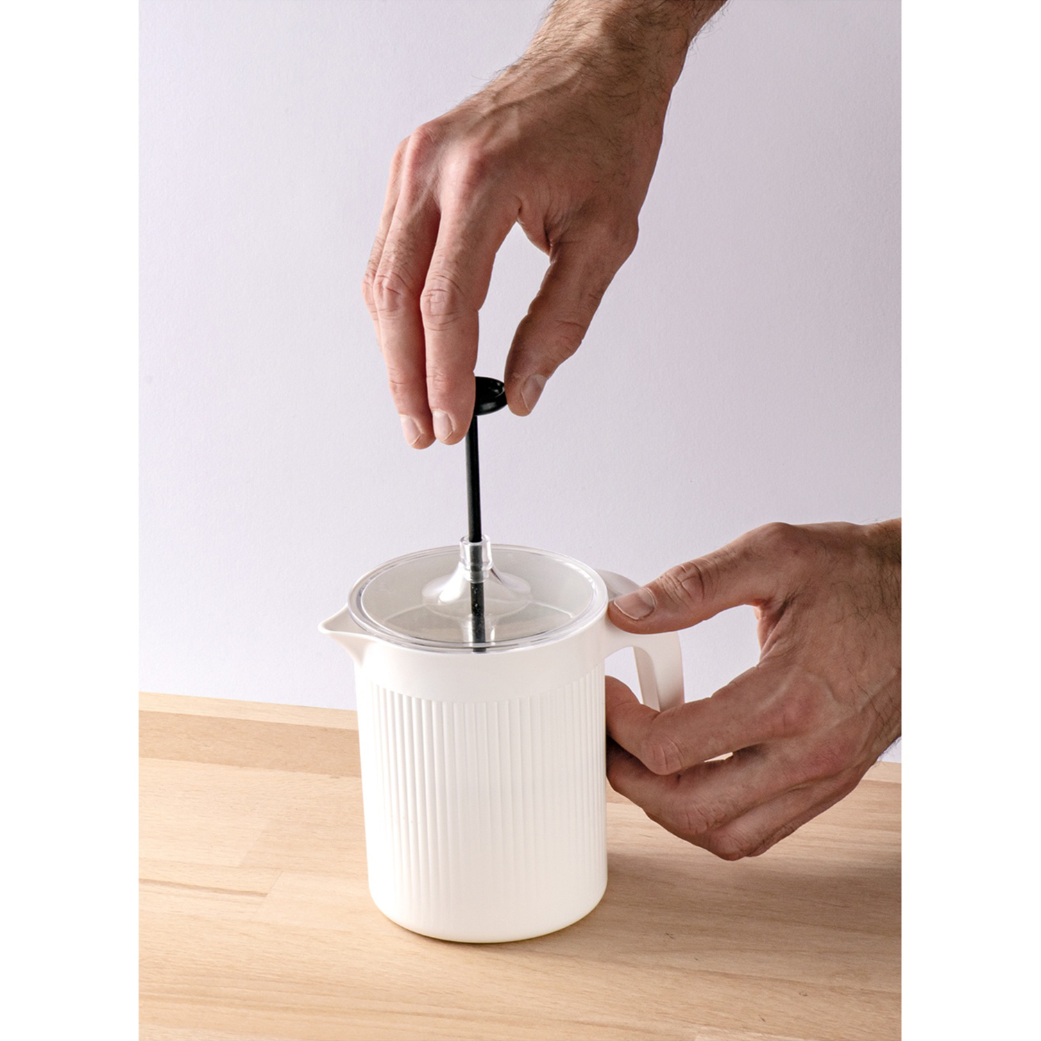 Una persona usa una caffettiera bianca. Le mani premono lo stantuffo. La caffettiera è su una superficie di legno, contro un muro bianco.