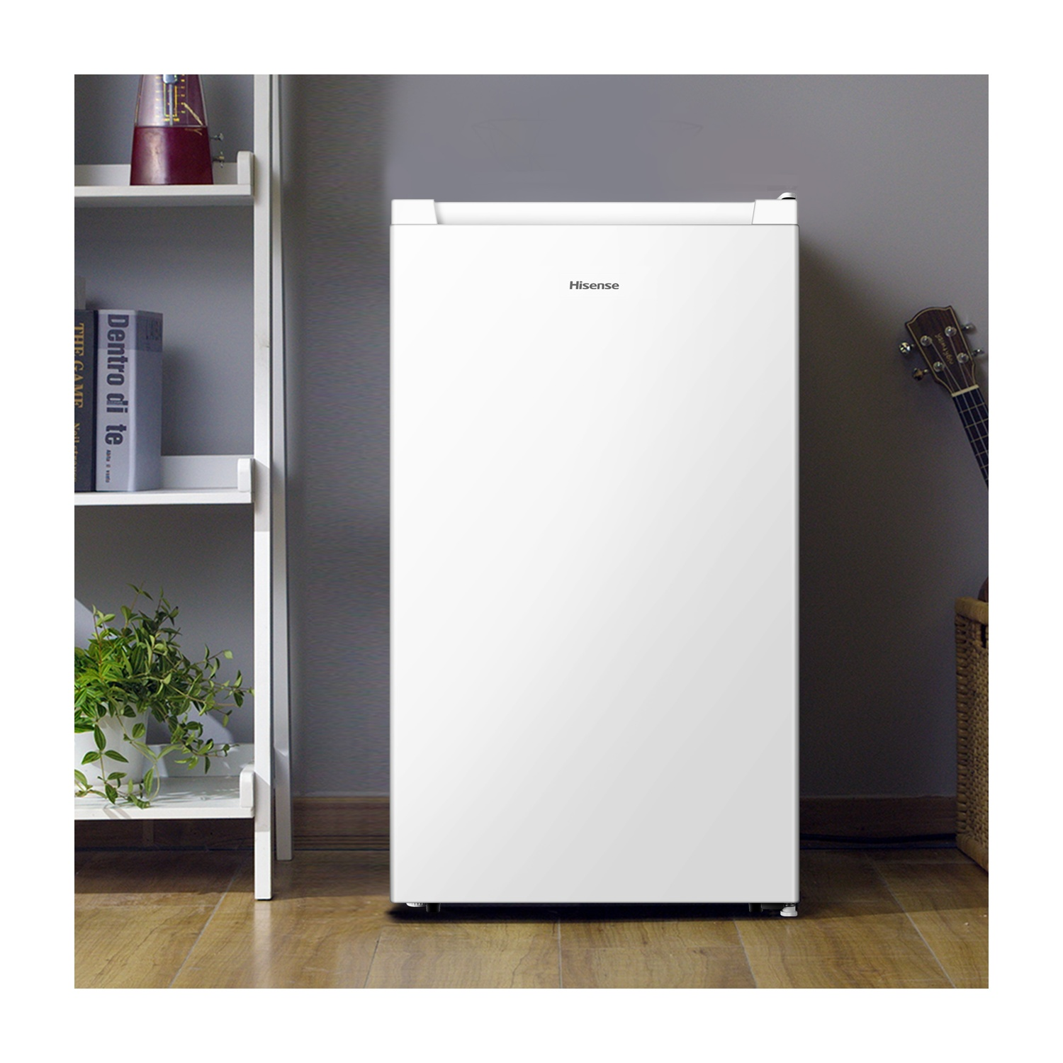 Un mini-frigo Hisense bianco contro un muro grigio. A sinistra, una mensola bianca con piante e libri. Una chitarra appoggiata al muro.