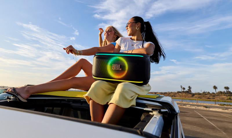 Zwei Frauen mit einem Lautsprecher auf einem Auto. Eine hält einen Lautsprecher. Klarer Himmel und Wasser im Hintergrund.