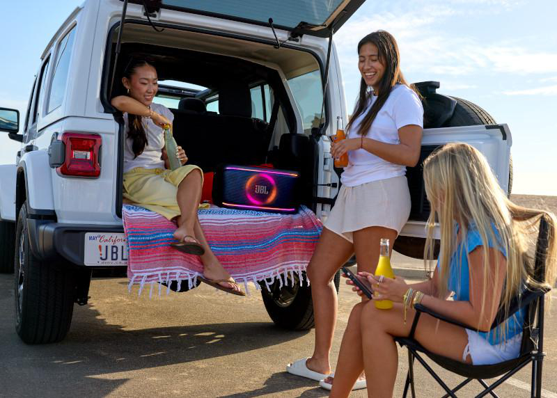Drei Frauen in der Nähe eines weißen SUVs mit offenem Kofferraum, einem Lautsprecher und Getränken. Eine sitzt drinnen, andere stehen und sitzen.