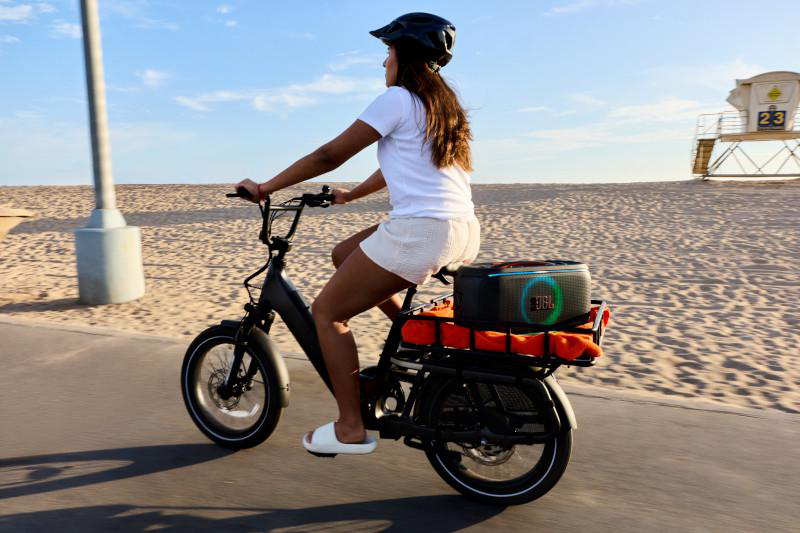 Eine Frau fährt mit einem schwarzen Fahrrad auf einem Weg, mit einem Lautsprecher auf dem Rücken. Strand und Rettungsschwimmerturm im Hintergrund.