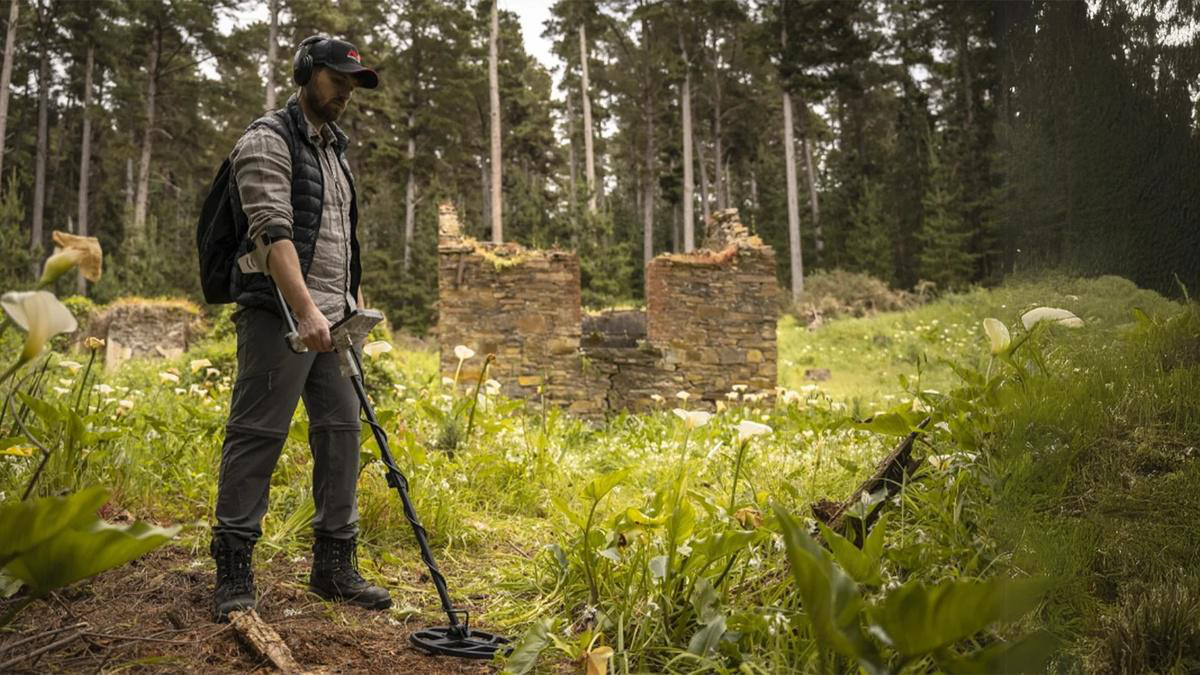 Mann mit Metalldetektor im Feld. Ruinen im Hintergrund. Weiße Blumen und hohe Bäume umgeben die Szene.