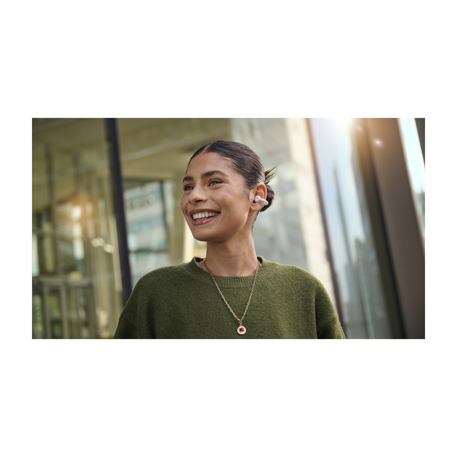Una donna sorridente con auricolari, maglione verde e collana. Lo sfondo mostra un edificio e la luce del sole.