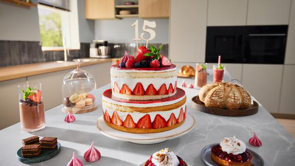 Ein zweistöckiger Kuchen mit Erdbeeren, Beeren und einem '15'-Topper. Andere Desserts und Brot liegen auf einem Tisch. Küche im Hintergrund.