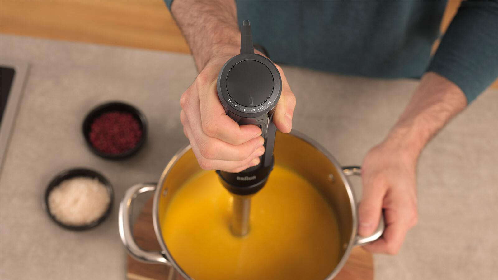 Eine Person verwendet einen Stabmixer in einem Topf mit gelber Suppe. Gewürze stehen in kleinen schwarzen Schalen auf der Theke.