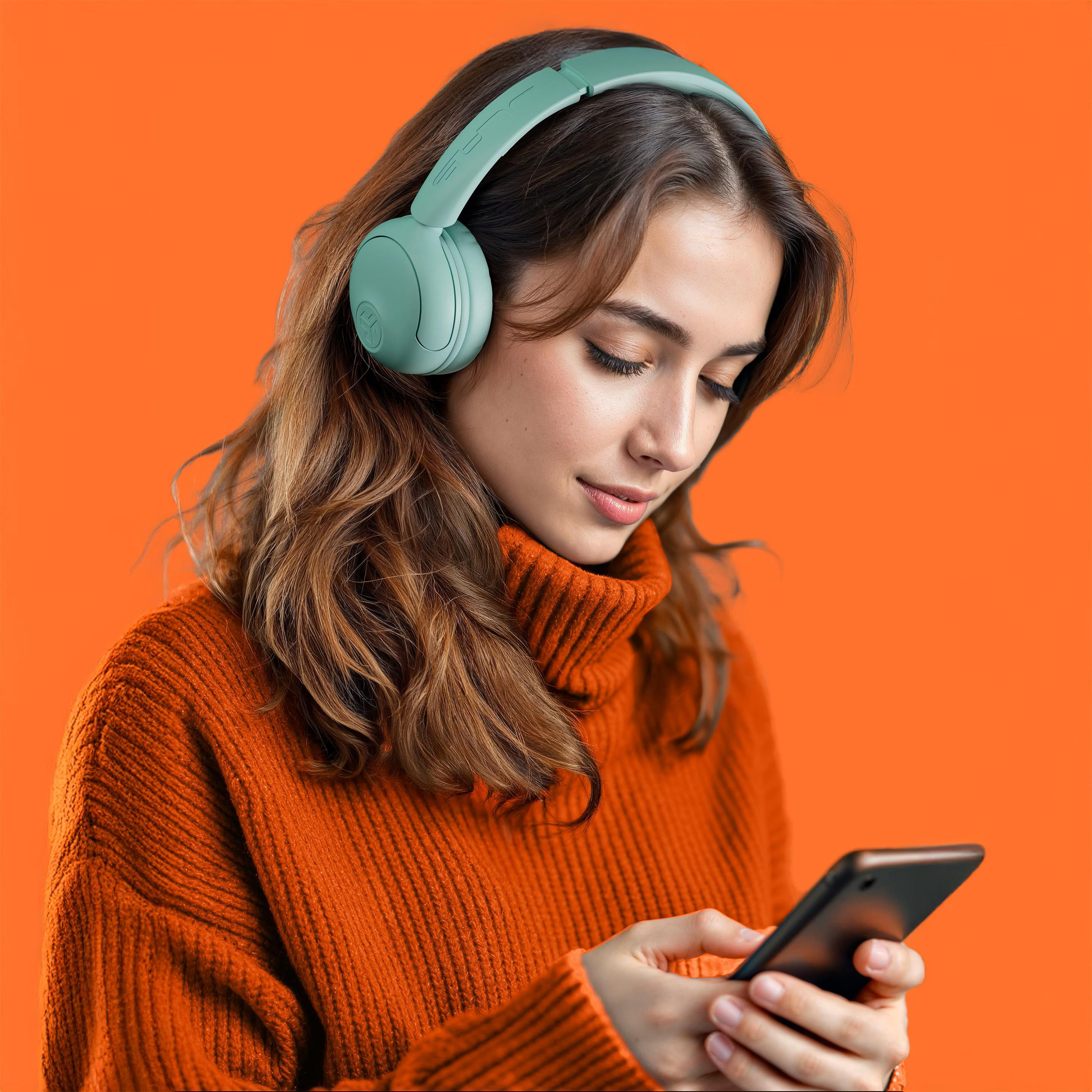 Eine Frau mit Kopfhörern und einem orangefarbenen Pullover schaut auf ein Smartphone vor orangefarbenem Hintergrund.