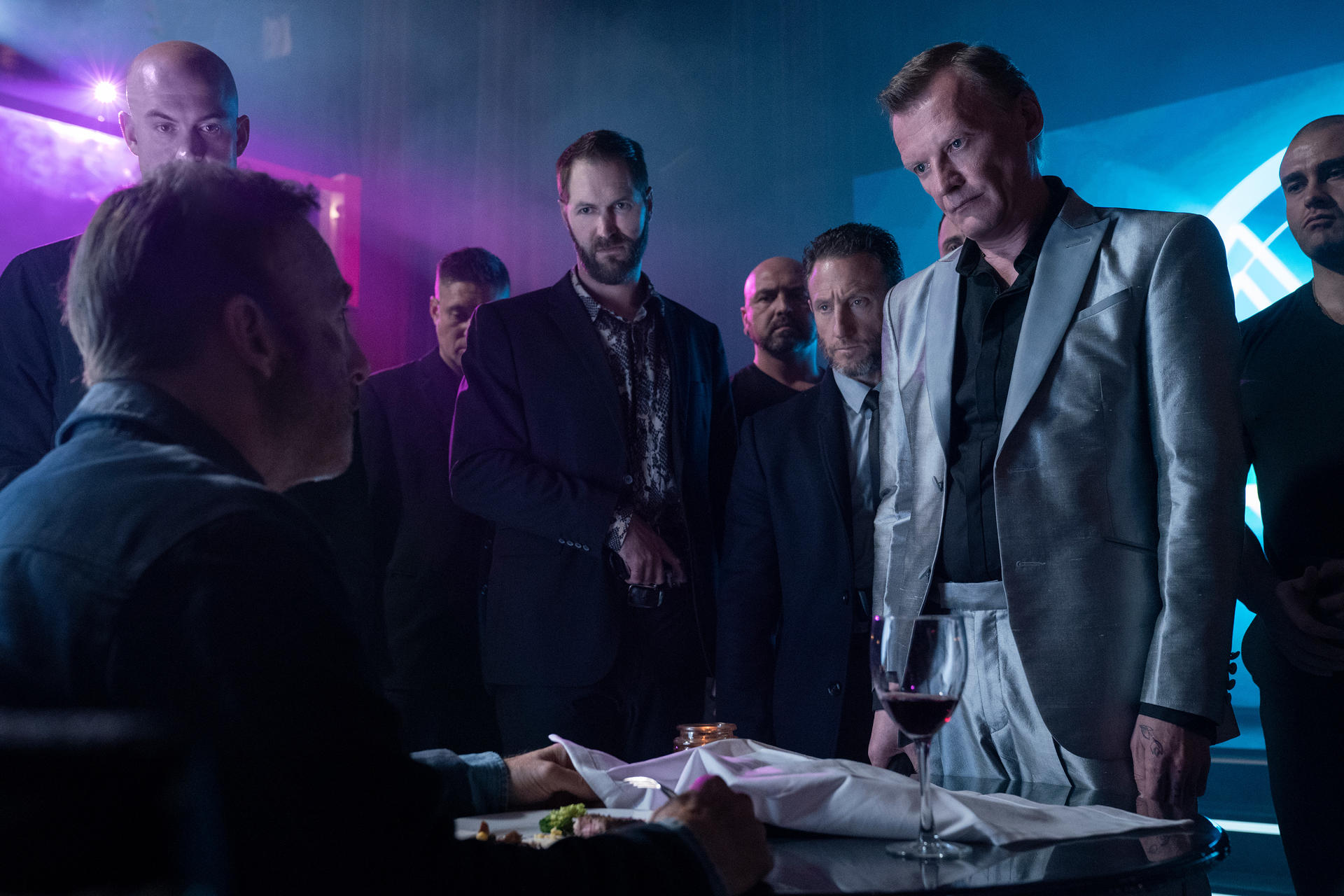 Des hommes en costume se rassemblent autour d'une table avec de la nourriture et du vin. Des hommes se tiennent derrière d'autres, dans un cadre sombre.