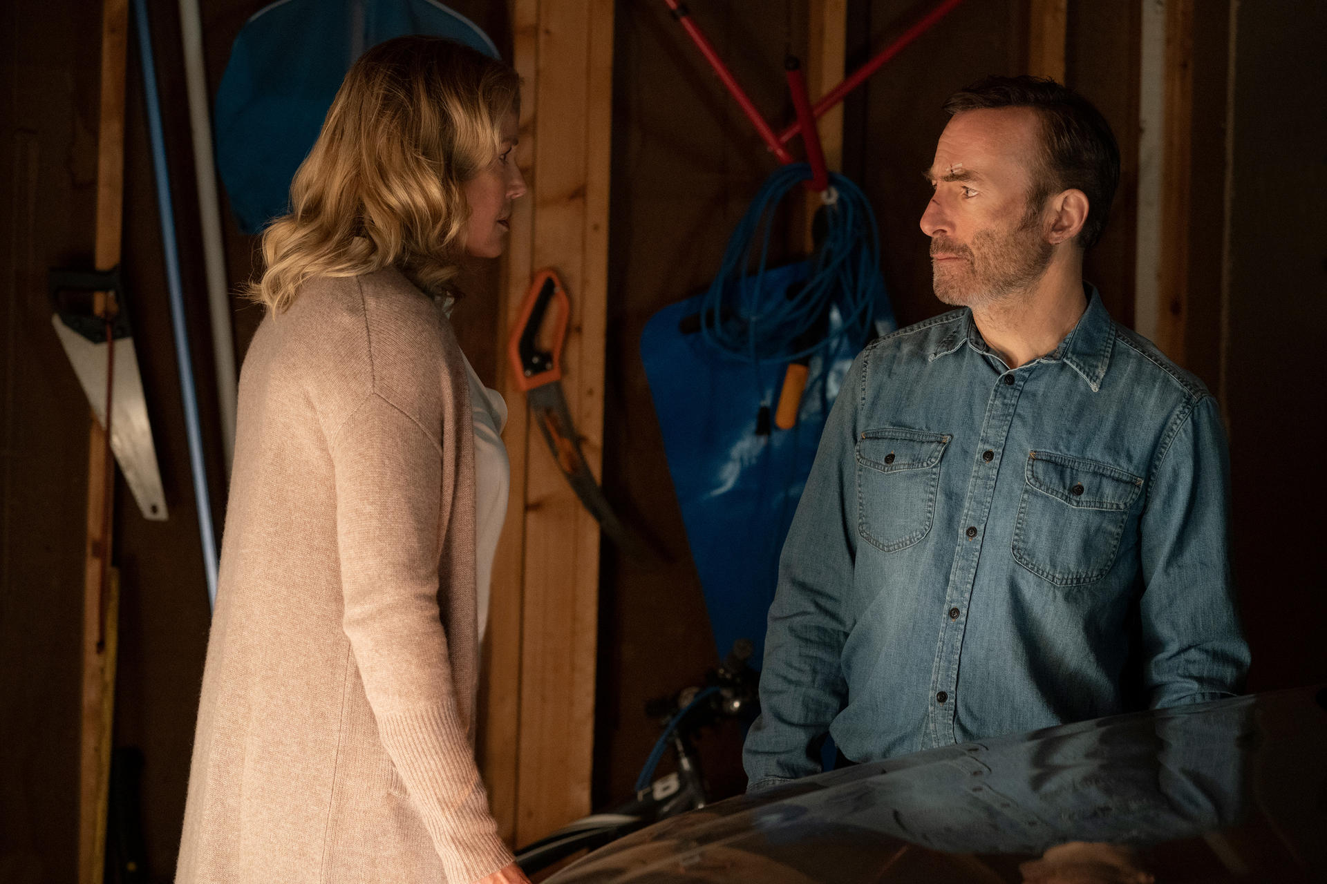Un homme et une femme se font face dans un garage. La femme porte un cardigan beige, l'homme une chemise en jean.