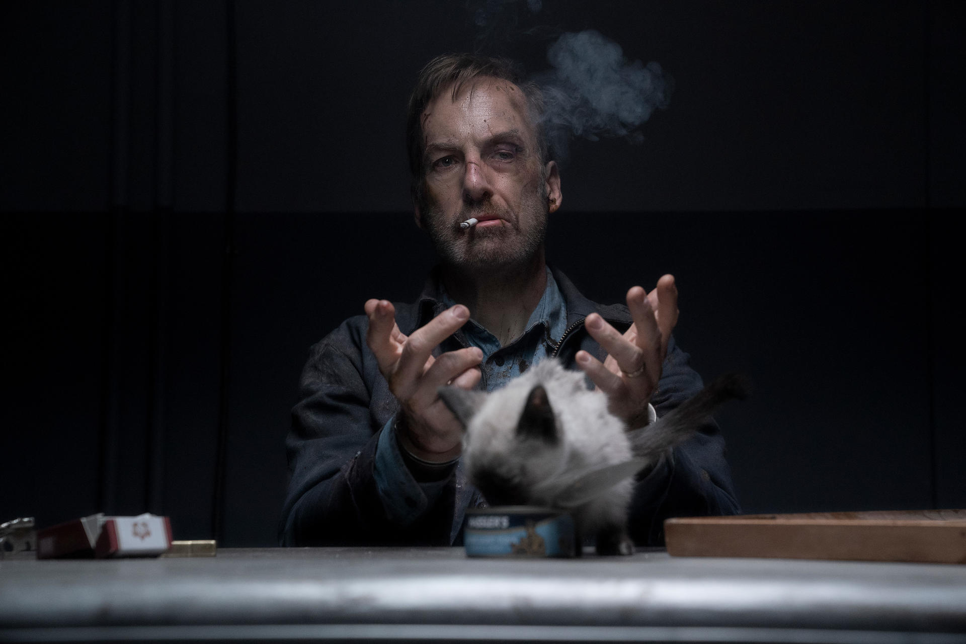 Homme avec cigarette, visage contusionné, gesticule vers un chat sur une table. Fumée monte. Fond sombre.