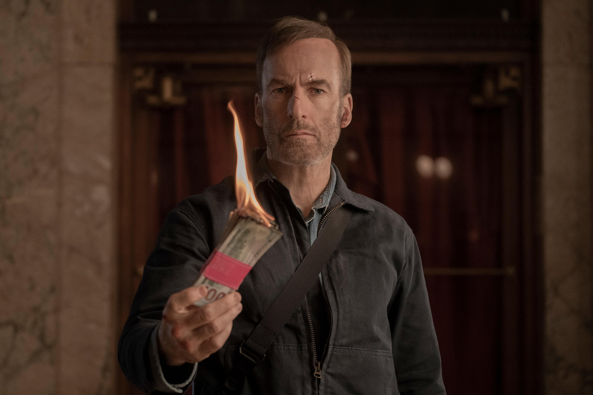 Un homme tient de l'argent en feu. Il porte une veste sombre et affiche une expression sérieuse. L'arrière-plan est une porte.