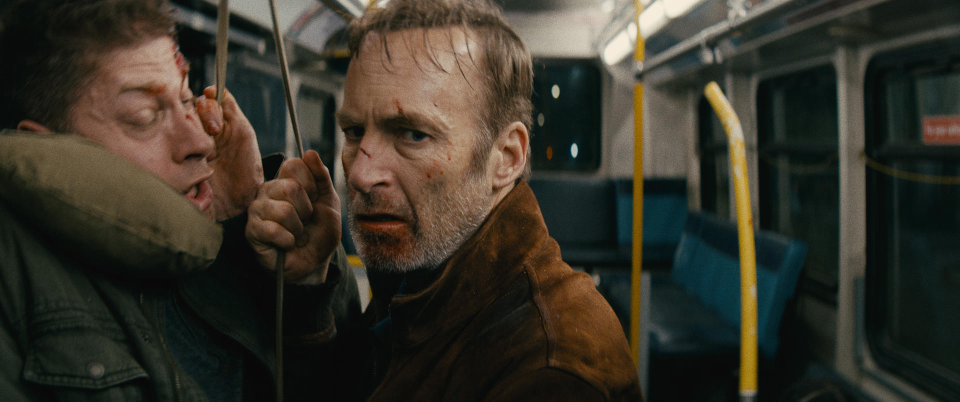 Deux hommes dans un bus, l'un avec le visage ensanglanté. L'autre homme regarde la caméra avec une expression déterminée. Intérieur du bus.
