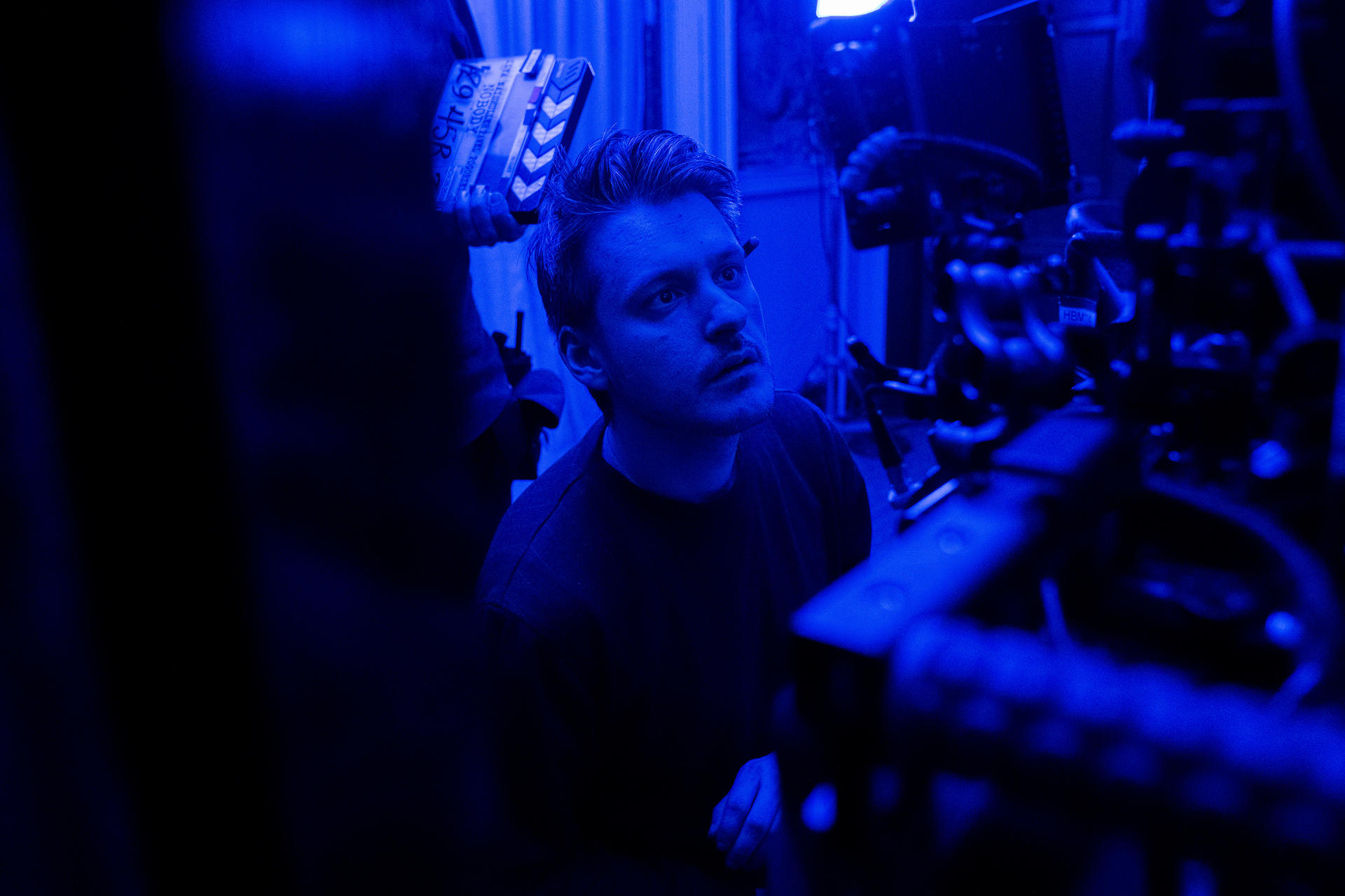 Un homme regarde vers le haut, avec un clap de cinéma en arrière-plan. La scène est éclairée en bleu, avec des caméras au premier plan.