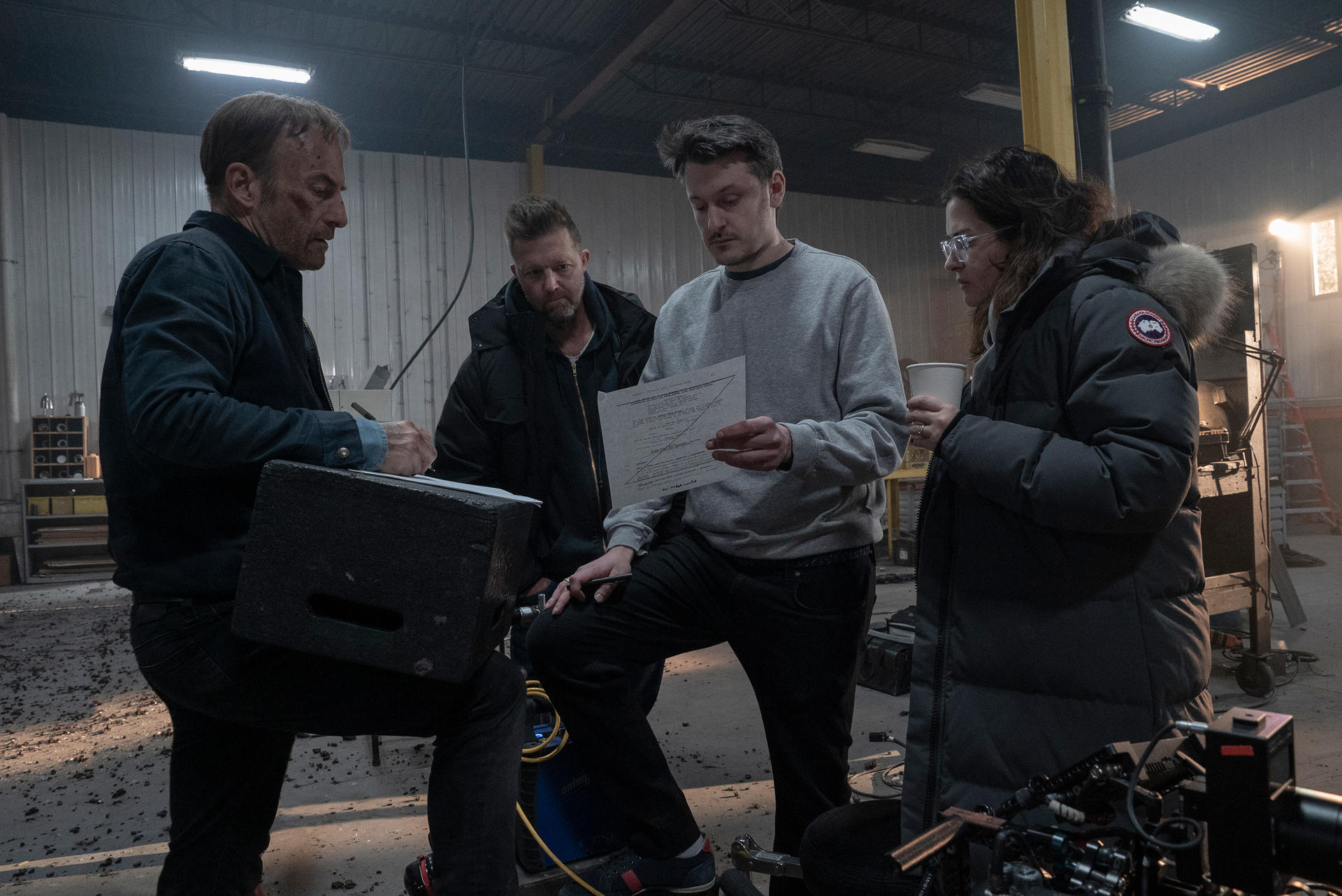 Quatre personnes sur un plateau de tournage, examinant des papiers et du matériel. L'un écrit sur un bloc, d'autres tiennent un papier et une tasse.