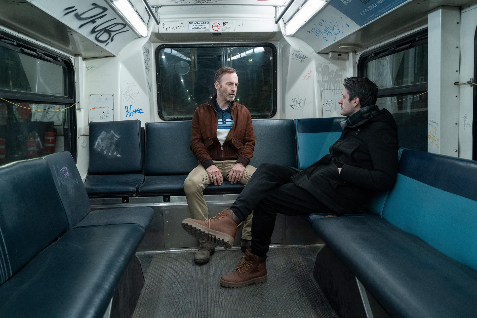 Deux hommes assis dans un métro. Graffiti sur les murs. L'un porte une veste marron, l'autre un manteau noir.