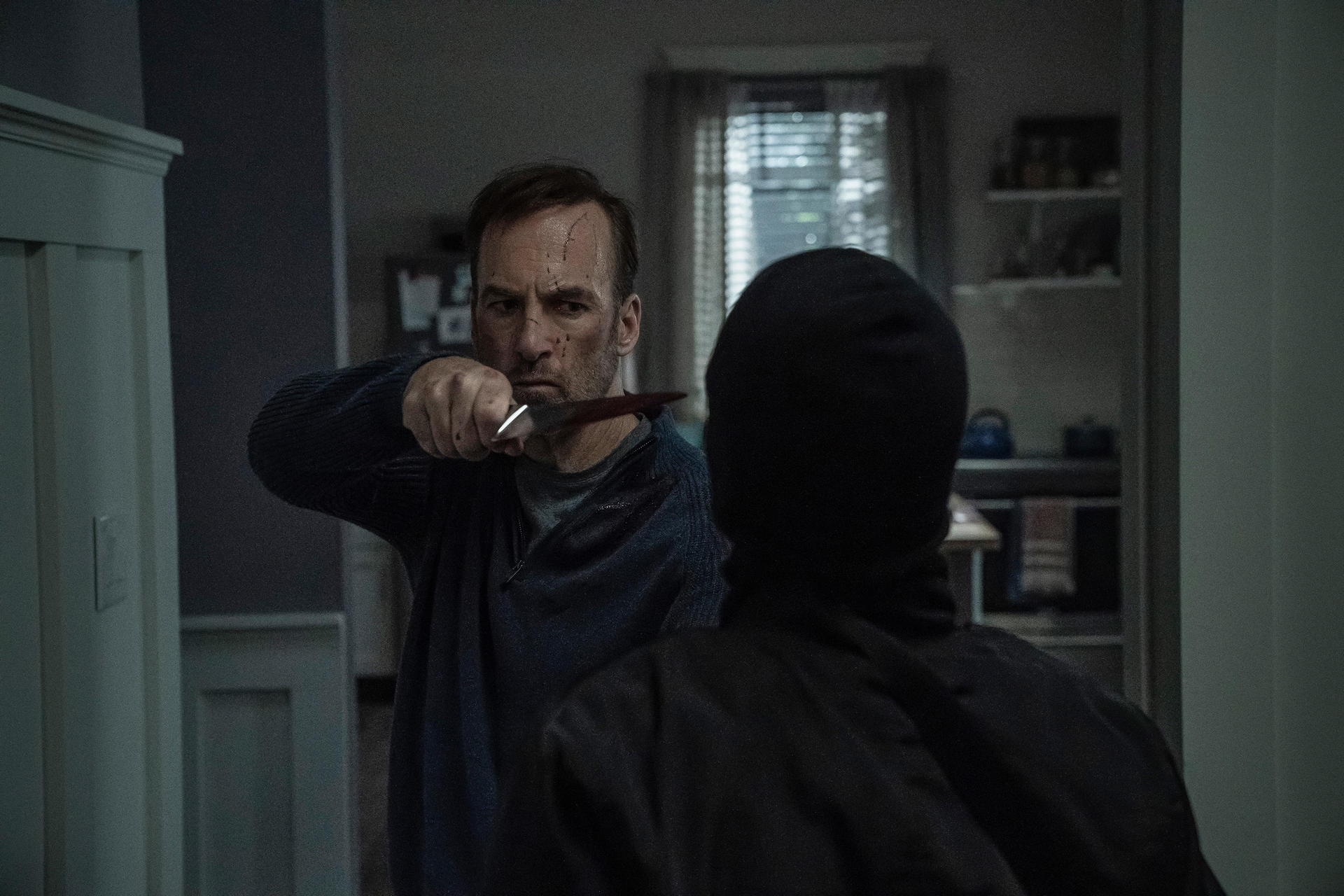 Un homme avec un couteau ensanglanté fait face à une personne masquée de noir. Scène intérieure, peut-être une maison. Ambiance tendue.
