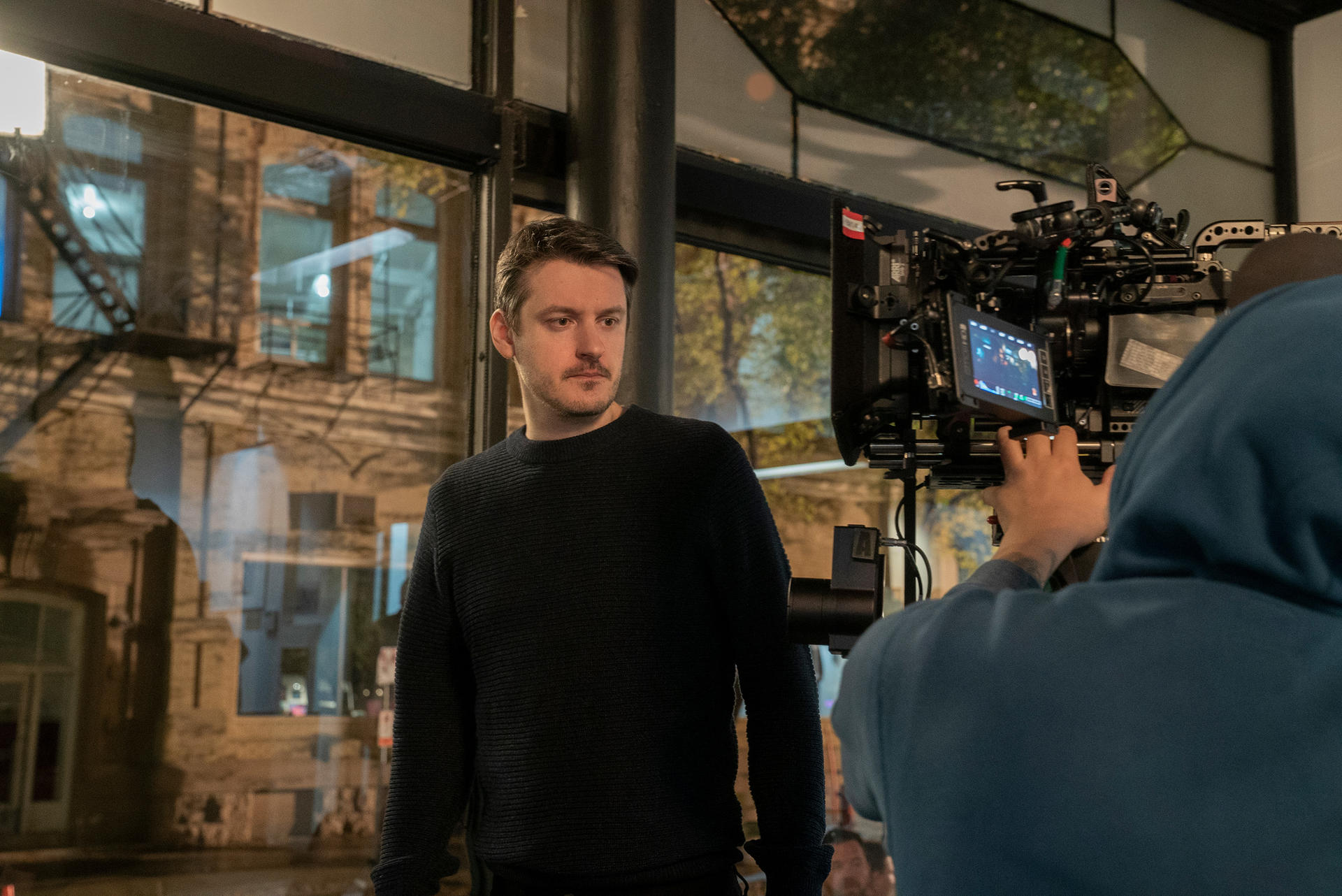 Un homme en pull noir regarde sur le côté. Derrière, une équipe de tournage utilise une caméra.