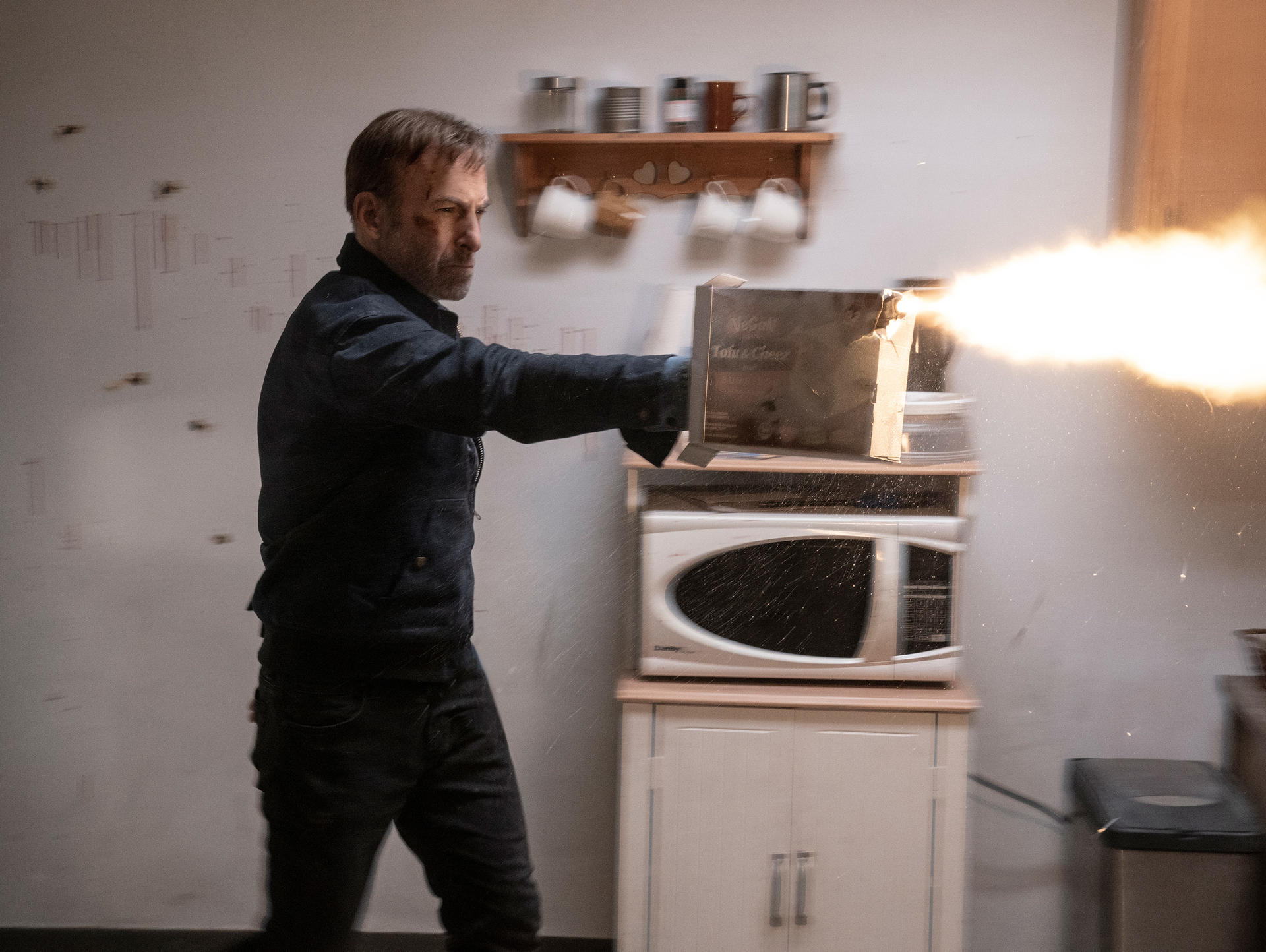 Un homme dans la cuisine tire avec une arme sur un micro-ondes. Des explosions et des débris remplissent l'air. Armoires et étagères de cuisine en arrière-plan.