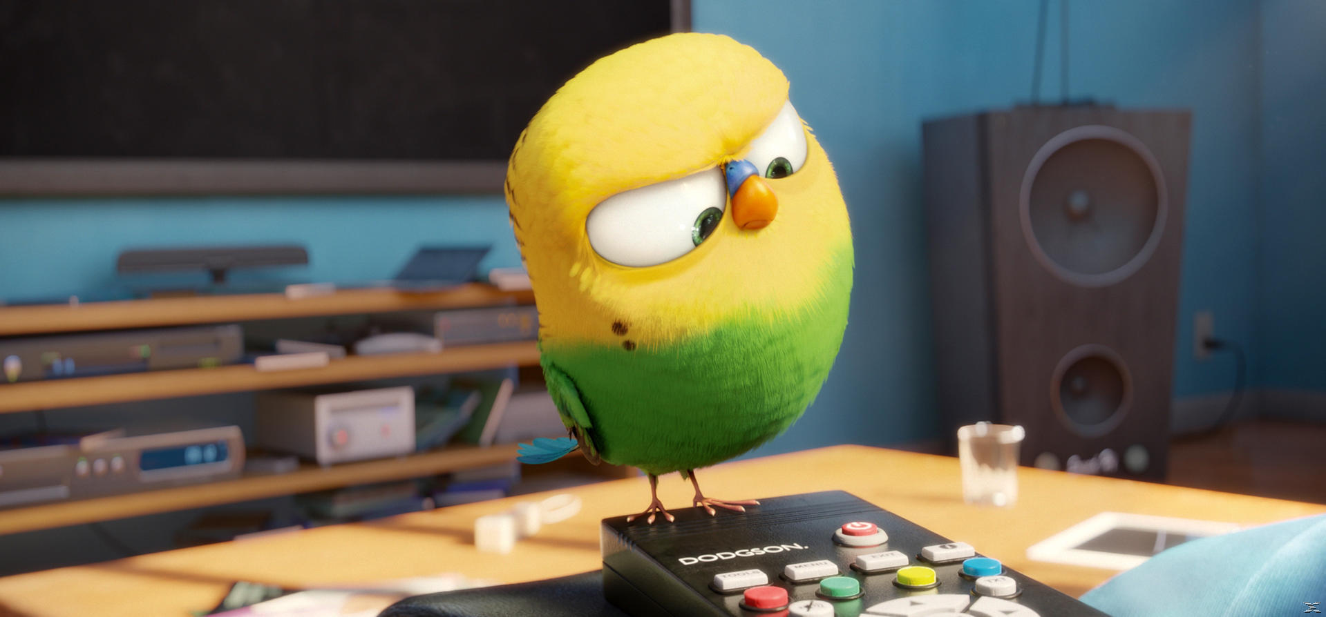 Une perruche de dessin animé est sur une télécommande. Elle est jaune et verte. Un haut-parleur et une télévision sont en arrière-plan.