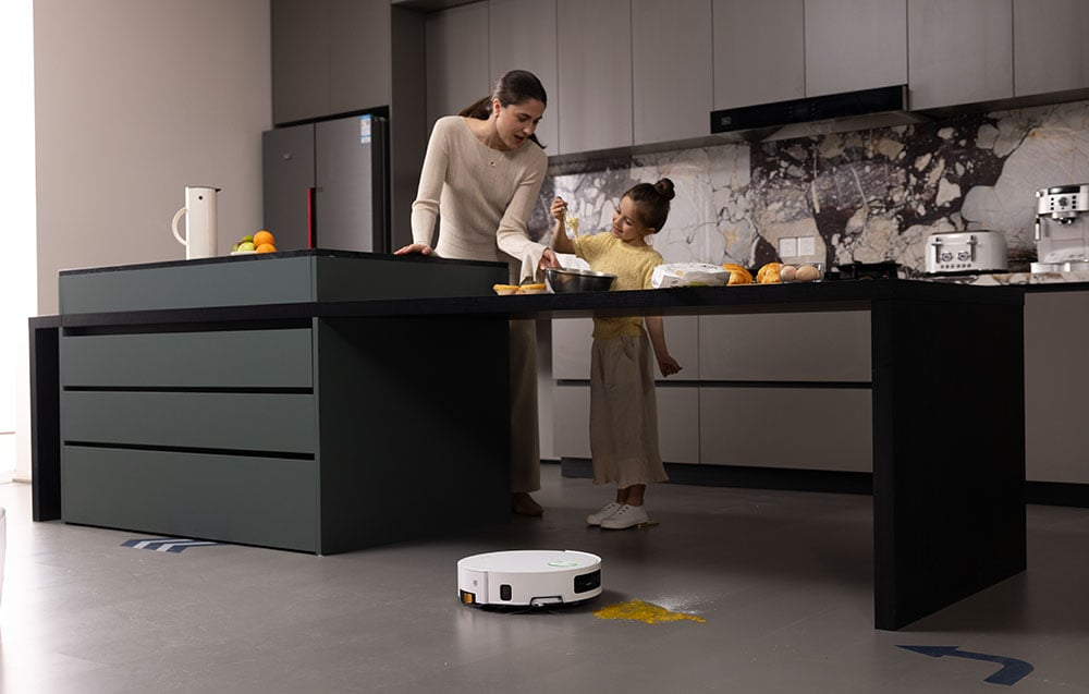 Egy nő és egy lány a konyhában főz. Egy robotporszívó tisztítja a padlót. Sárga anyag van a padlón szétszórva.