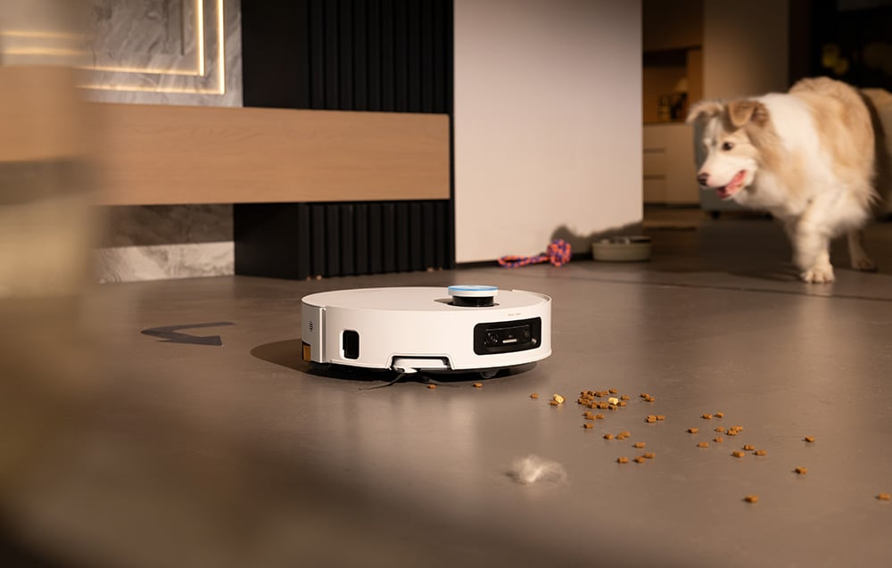 Fehér robotporszívó szürke padlón. Egy kutya a háttérben. Szétszórt étel és szőr a padlón.
