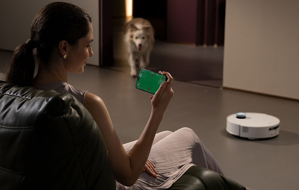 Nő zöld képernyős telefonnal, robotporszívót néz. Egy kutya sétál a háttérben.