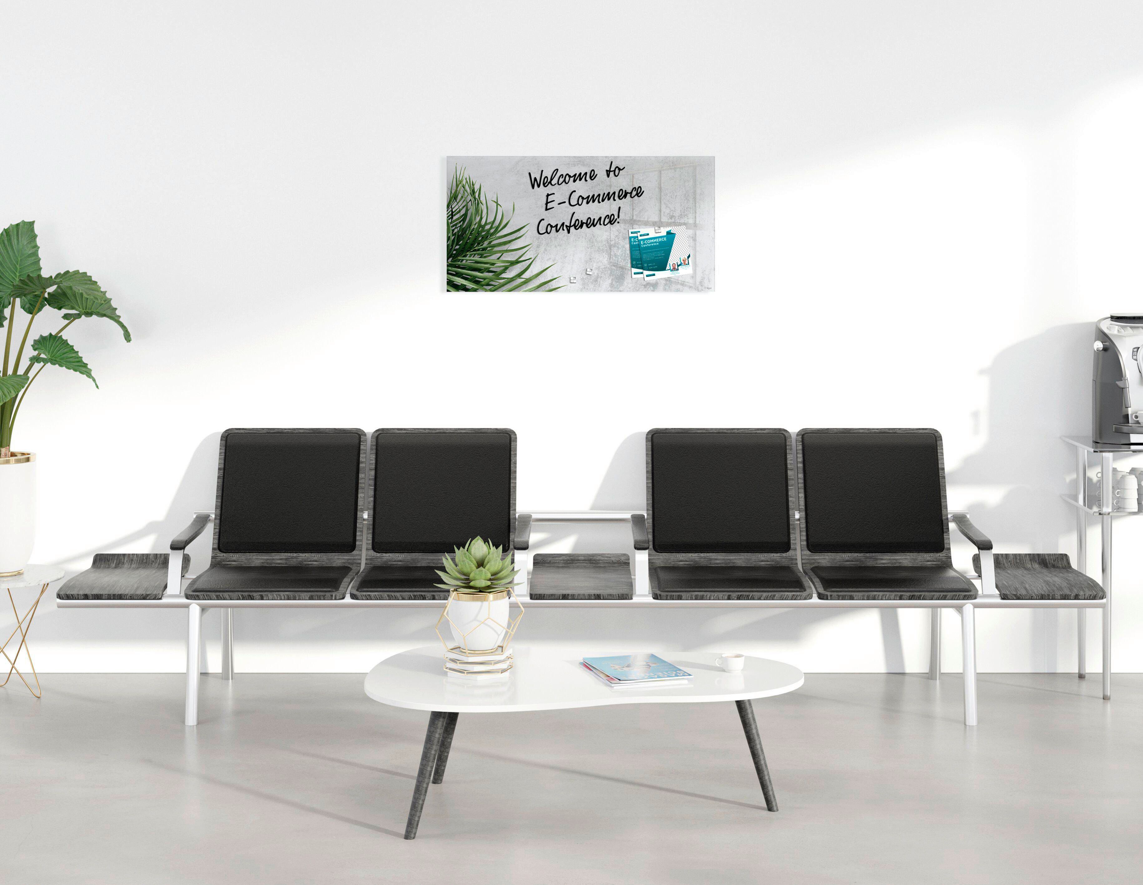 Wartebereich mit Stühlen, Couchtisch und Schild für E-Commerce-Konferenz. Pflanze und Kaffeemaschine vorhanden.