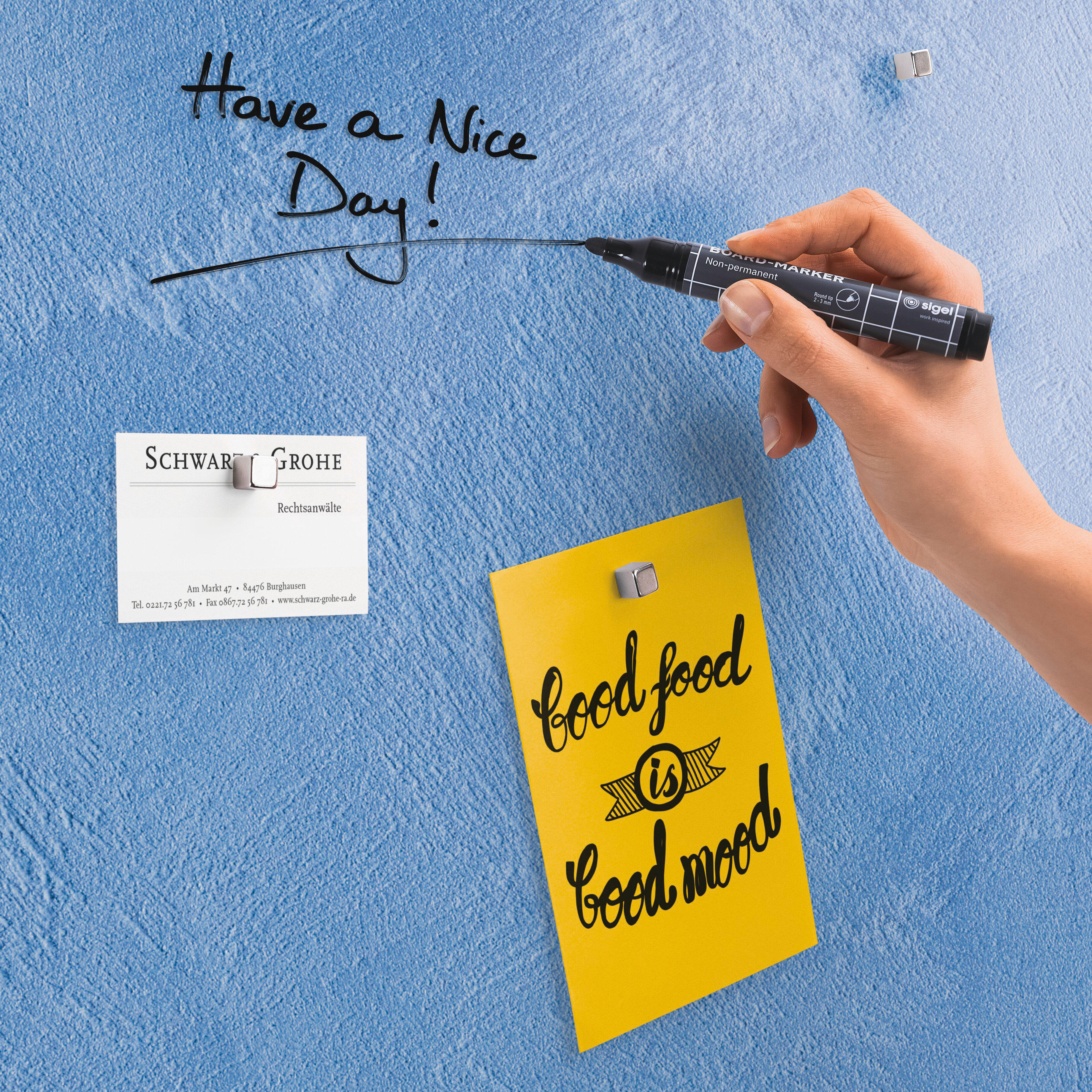 Eine Hand, die einen Marker hält und auf eine blaue Wand schreibt. Notizen sind mit Magneten befestigt, darunter eine gelbe mit der Aufschrift 'gutes Essen ist gute Laune'.