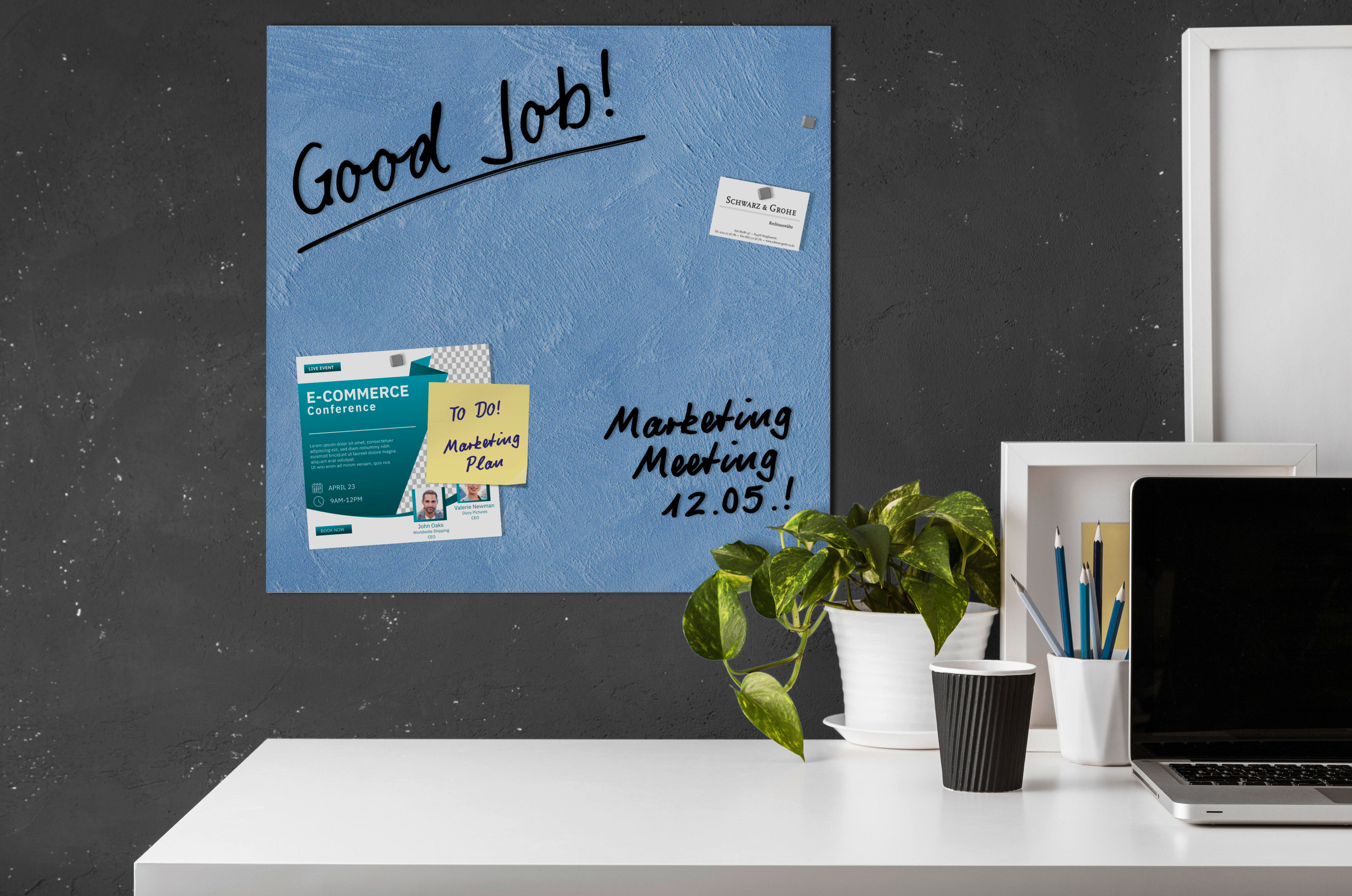 Blaue Tafel mit Text, Notizen und einer Pflanze. Ein Laptop und Bleistifte stehen auf dem weißen Schreibtisch.