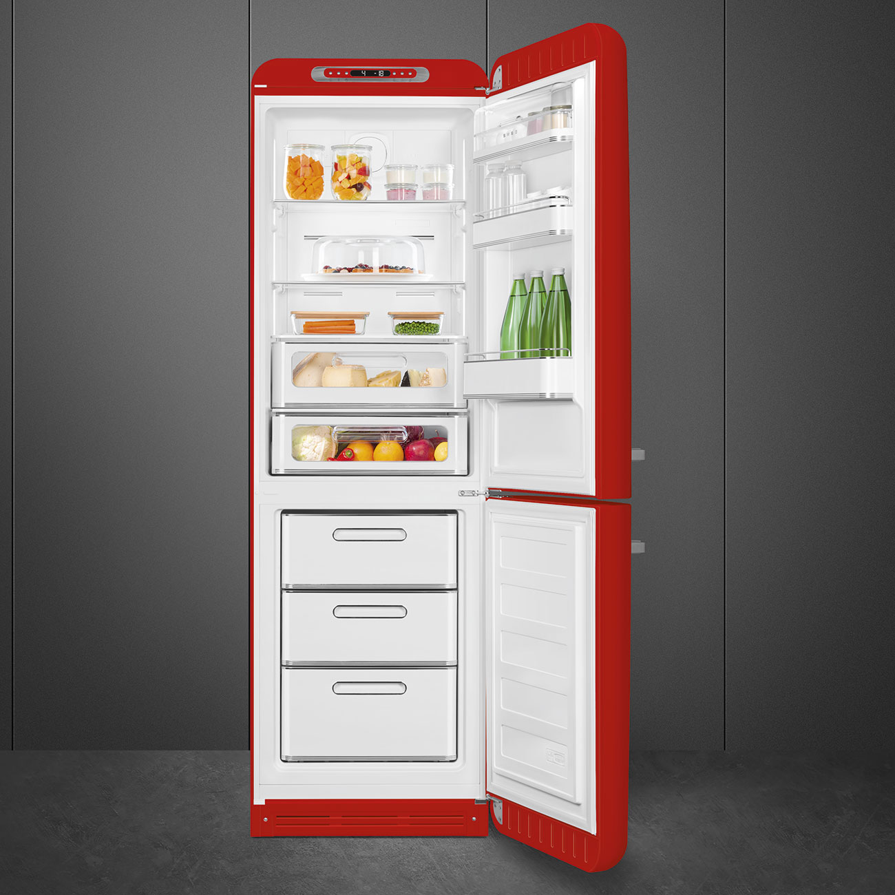 Offener roter Kühlschrank mit Lebensmitteln. Enthält Gläser, Gemüse, Käse, Früchte und Flaschen. Grauer Wandhintergrund.