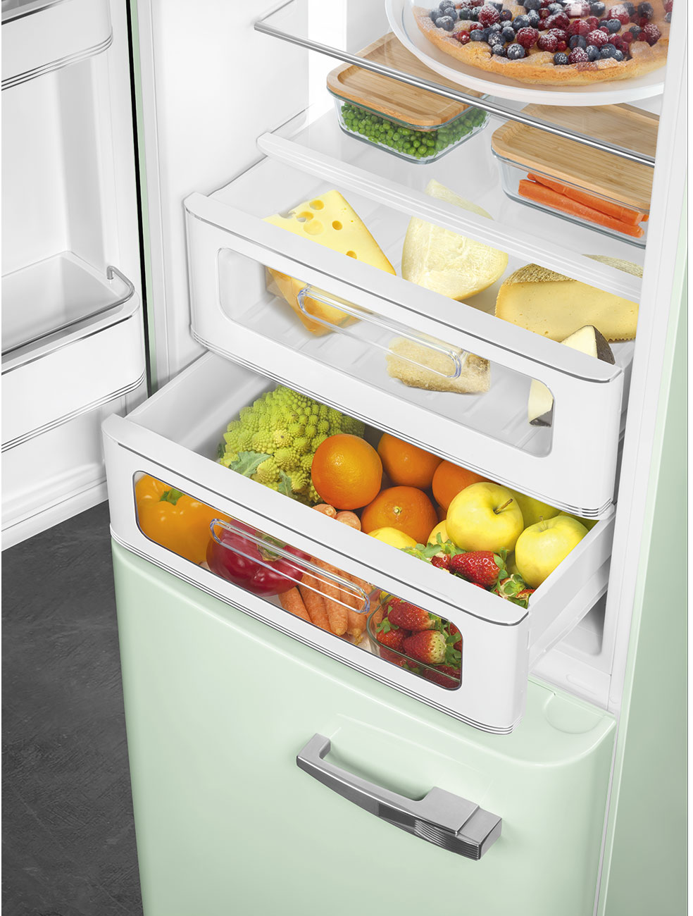 Im Kühlschrank befinden sich Schubladen mit Essen: Käse, Orangen, Äpfel und Erdbeeren. Auf dem obersten Regal befindet sich ein Kuchen. Der Kühlschrank ist mintgrün.
