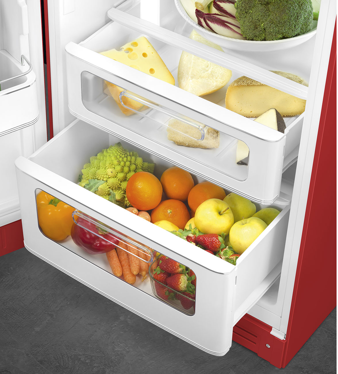 In einem Kühlschrank befinden sich Schubladen mit Käse, Orangen, Äpfeln, Erdbeeren und Gemüse. Rotes und weißes Innere.