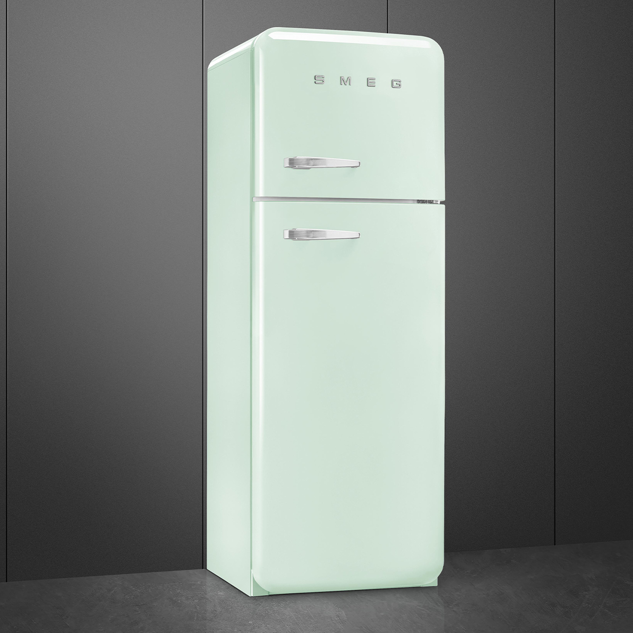Ein mintgrüner Smeg-Kühlschrank. Er hat zwei Türen und Chromgriffe und steht vor einer dunkelgrauen Wand auf einem grauen Boden.